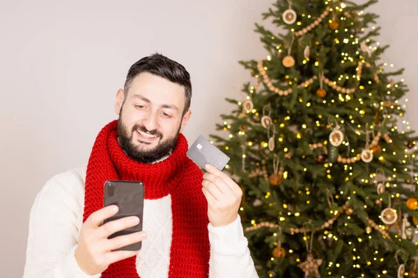 Glücklicher brünetter bärtiger Mann, der online per Kreditkarte für Weihnachtsgeschenke bezahlt, das Smartphone in der Hand hält, auf den Bildschirm schaut, zu Hause neben dem Weihnachtsbaum steht und ein rotes Tuch trägt. Online einkaufen. — Stockfoto