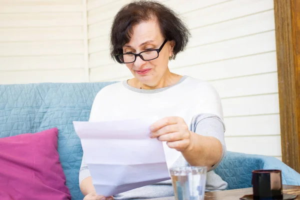 Удивлённая счастливая пожилая женщина в очках, читающая бумажный документ или почтовое письмо от друга, сидящая на диване на домашней террасе. Отказ от приятной бумажной работы банковское уведомление или почтовая переписка — стоковое фото