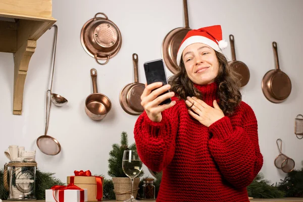 Genç ve mutlu esmer kadın Noel Baba şapkası takıyor ve evde cep telefonu kullanıyor. Yılbaşı partisinde cep telefonuyla video görüşmesi yapmak, uygulama kullanarak uzaktan arkadaşlarla ve ailelerle sohbet etmek.