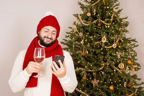 Uśmiechnięty brodaty mężczyzna wykonujący wideo połączenie ze smartfonem, noszący czerwony kapelusz i szalik, stojący w pobliżu choinki. Najlepsze przyjaciółki pijące szampana. Wideokonferencja internetowa — Zdjęcie stockowe