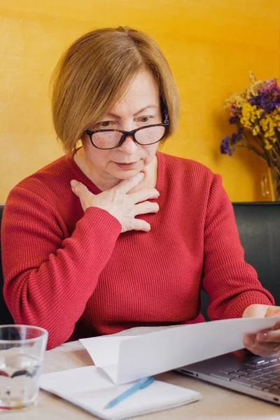 年长的女人在阅读纸质文件或信件时，对坐在家里的意想不到的新闻感到惊讶和震惊。收到银行账户余额或坏账。 免版税图库照片