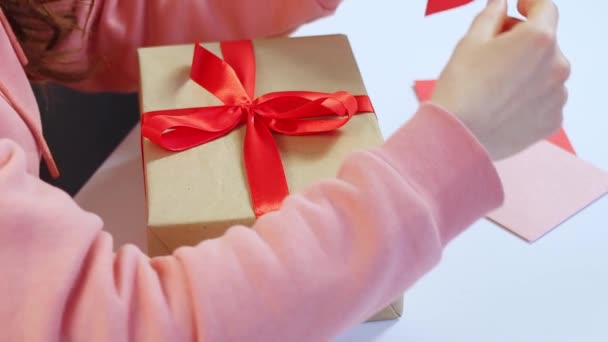 Sevgililer günü kartpostalını kırmızı kalp şeklinde koyan, kırmızı kurdeleli hediye paketi hazırlayan ve bayram ya da doğum günü için eğilen genç bir kadın. Aşk ilanı, ilişki kavramı. Yüksek kaliteli FullHD görüntüler — Stok video