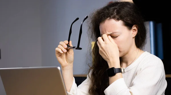 Tired young woman after computer work, keeping eyes closed and massaging nose, taking off glasses. Overworked female freelancer suffering from blurry vision after long laptop use, eye strain problem