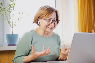 Happy mature woman in eyeglasses talking using video conference call application on laptop, sitting at home.Tutoring,holding online call with client, webinar, educational class lecture using internet