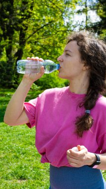 Eğitimden sonra açık yeşil park bahçesinde duran plastik şişeden temiz maden suyu içen genç bir kadın. Sıcak yaz gününde su dengesini sağlamak. Susuzluk giderici. Dikey