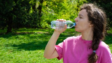 Eğitimden sonra açık yeşil park bahçesinde duran plastik şişeden temiz maden suyu içen genç bir kadın. Sıcak yaz gününde su dengesini sağlamak. Susuzluk giderici. Boşluğu kopyala