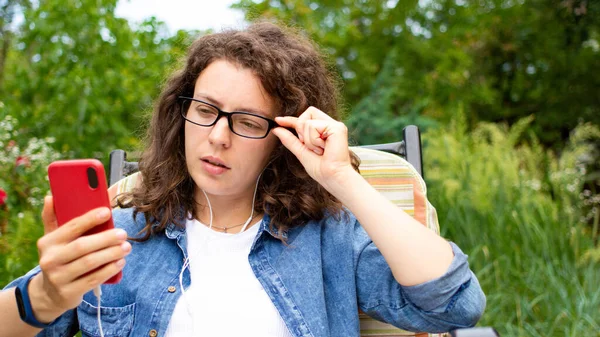 Kulaklıklı ve gözlüklü genç bayan öğrenci yeşil bahçede oturmuş akıllı telefon ekranına bakıyor. Çevrimiçi Webinar 'ı izliyor, dil öğreniyor, ses kursunda 5 g internet kullanıyor.