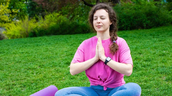 Calm young woman with closed eyes sitting in lotus pose, practicing yoga breathing techniques on green grass in garden outdoor,wearing pink t-shirt.Meditation and pranayama.Mental health care concept Лицензионные Стоковые Изображения
