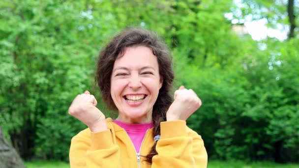 Smiling overjoyed beautiful brunette young woman in yellow hoodie posing doing winner gesture, say Yes standing in green garden outdoors. People sincere emotions, lifestyle concept. Slow motion — Video