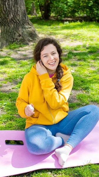 Smiling young woman student wearing wireless headphones looking at camera, wearing yellow hoodie and blue leggings, sitting on green grass in park outdoors. Preparing use modern device. Vertical
