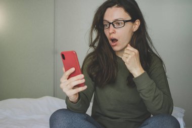 Shocked young brunette woman looking at mobile phone, sitting in bed at home, reading disturbing news in media, watching video content, feeling frustrated by unexpected information.Received bad email
