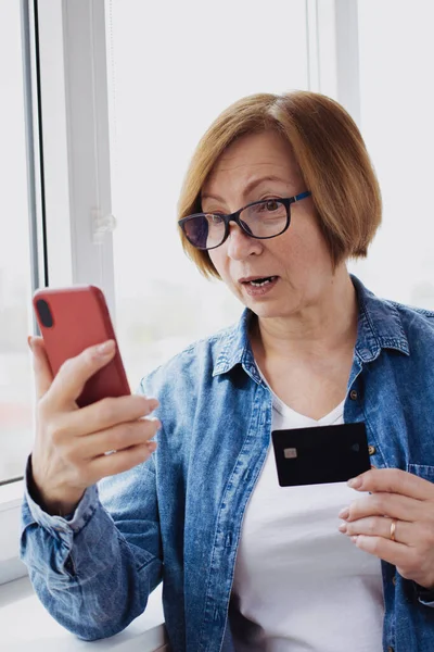 Disappointed elderly woman holding bank credit card, looking confused on mobile phone standing near window indoor. Senior online buyer has debt problems, failed transaction, money error. Vertical. — 스톡 사진