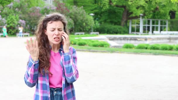 Furious frustrated yelling aggressive young curly woman talking on mobile phone, standing in park outdoors, actively gesticulates.Conflict conversation, explaining something. High quality 4k footage — Vídeo de stock