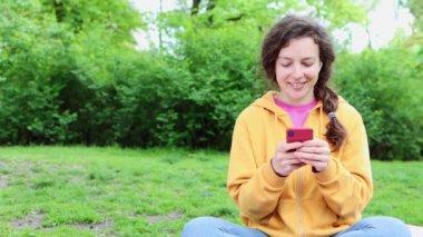 Yeşil şehir parkında oturan genç ve mutlu bir kız öğrenci cep telefonu ekranına bakıyor. Kısa mesaj, internette gezinme, sosyal medyada 5g internet bağlantısı kullanarak sohbet. Boşluğu kopyala.