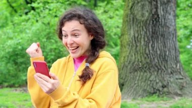 Çevrimiçi zaferi kutlayan genç kıvırcık kadın dışarıda yeşil bahçede oturuyor, cep telefonunu tutuyor e-postalarını kontrol ediyor, heyecandan uçuyor. Kazanan başarı anının tadını çıkarır, bahis konsepti. Yavaş çekim