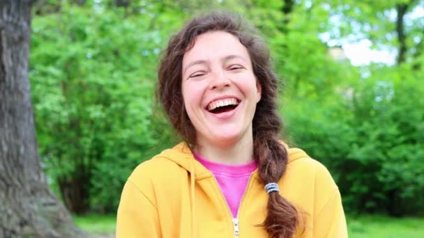 Cheerful casual young woman student laughing out loud at humorous joke looking at camera standing in green garden outdoors, with funny face enjoy sincere positive emotion, wearing yellow hoodie. — Stockvideo