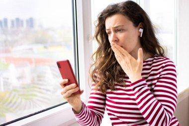 Şok olmuş genç bir kadın cep telefonuna bakıyor, pencerenin yanında duruyor. Medyada rahatsız edici haberler okumak, video içeriği izlemek, beklenmedik bilgilerle hayal kırıklığına uğramak. Alınan kötü e- posta