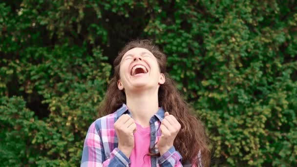 Curly brunette young woman bursting with laughter at humorous joke look at camera standing outdoors in green garden, with funny face enjoy sincere positive emotion,wearing checkered shirt.Slow motion — ストック動画