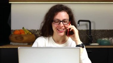 Smiling young business woman professional talking on phone using laptop sit in kitchen at home. Customer make mobile call confirming online website shopping order delivery concept. Slow motion