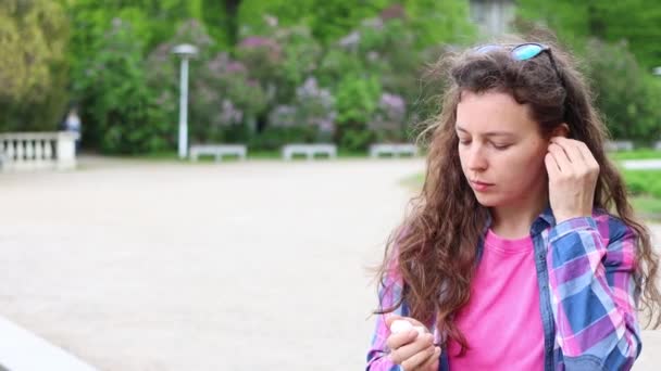 Young beautiful curly woman takes small wireless earbuds from charging case and puts headset into ears, standing in park outdoors. Listen to music using smartphone. High quality 4k footage. — Vídeo de stock