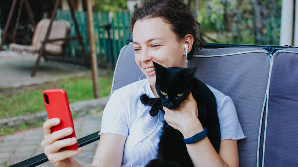 Cheerful brunette girl using smartphone and hugging black cat sitting on sofa at home terrace outdoors. Video call, virtual online conference using wireless internet connection, distant communication