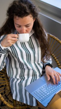 Serbest çalışan kıvırcık kadın kahve içiyor, dizüstü bilgisayar kullanıyor, kafedeki rattan koltukta oturuyor. İnternette uzaktan sohbet ve internet sitesi için 5g internet bağlantısını kullanarak çalışıyor. Dikey