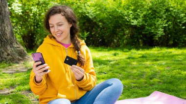 Mutlu genç bayan müşteri ya da alıcı kredi kartıyla akıllı telefondan alışveriş yapıyor, bahçedeki yeşil çimlerin üzerinde oturuyor. İnternet ödemesi yapmak, online alışveriş, güvenli bankacılık kavramı.