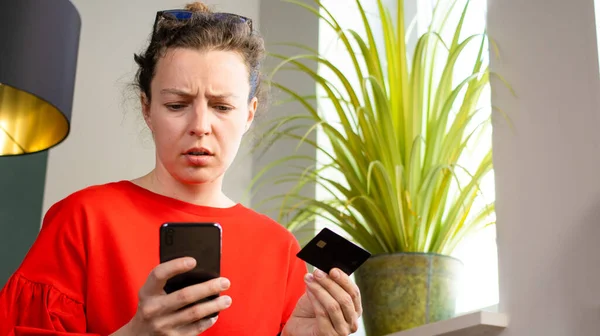 Confused brunette girl holding bank credit card, looking at mobile phone sitting indoors in modern interior near green plant. Debt problems, failed transaction, money error, contacting with scammer