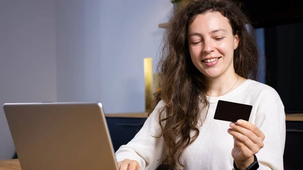 快乐的年轻女子在家里拿着银行卡，坐在厨房里用笔记本电脑模拟。上网、网上购物、预订旅馆、订票、定单、打车、用银行捐款 图库图片