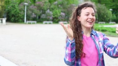 Neşeli, kıvırcık, gülümseyen, el sallayan, cep telefonuyla video görüşmesi yapan, şehir parkında açık havada duran, sanal uygulamayla sohbet eden, 5G internet bağlantısı kullanan genç bir kadın. 4k görüntü.