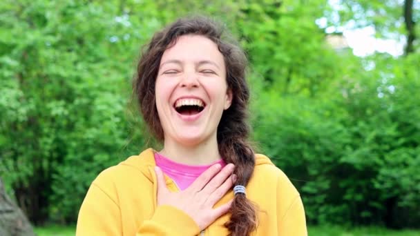 Χαρούμενη περιστασιακή νεαρή γυναίκα γελάει δυνατά σε χιουμοριστικό αστείο κοιτάζοντας κάμερα στέκεται στο πράσινο κήπο σε εξωτερικούς χώρους, με αστείο πρόσωπο απολαμβάνουν ειλικρινή θετική συγκίνηση, φορώντας κίτρινη κουκούλα. Αργή κίνηση — Αρχείο Βίντεο