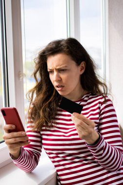 Kafası karışmış internet alıcısı bin yıllık kadın kredi kartı tutuyor, cep telefonuna bakıyor, pencerenin yanında duruyor. Borç problemleri, güvensiz çevrimiçi ödeme, başarısız işlem, hesapta yeterli para yok.