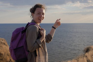 Heyecanlı yürüyüşçü kız deniz kenarındaki dağ zirvesinde uzun bir yürüyüşten sonra parmağını okyanusa doğrultarak şaşırmış bir yüz ifadesiyle kameraya bakıyor. Arka planda gün batımı ışığı ve bulutlu gökyüzü. Boşluğu kopyala