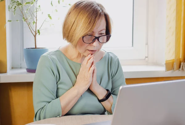 穿着眼镜的成熟女人惊讶地看着笔记本电脑 在媒体上阅读令人不安的新闻 观看视频内容 坐在室内 感受希望 祈祷幸福的结局 收到不良电子邮件 图库图片