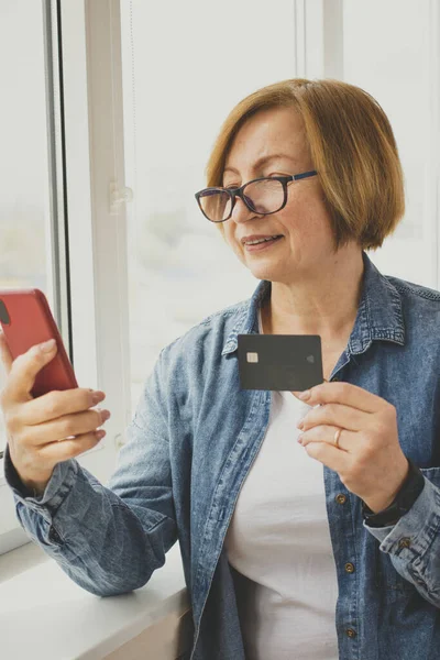 Akıllı telefon kullanarak banka kartı tutan, internet ödemesi yapan, internetten alışveriş yapan, otel kiralayan, uygulamayla taksi çağıran, kapalı bir pencere kenarında duran mutlu yaşlı bir kadın. Çevrimiçi bankacılık. Dikey. Model