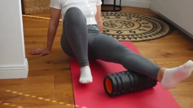 Spor kadın ısınmak için köpük silindiri üzerinde egzersiz yapıyor, modern oturma odasındaki pembe paspasın üzerinde masaj yapıyor. Evde bacağındaki ağrıyı gidermek için egzersiz. Spor tıbbında rehabilitasyon. 