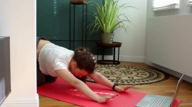 Genç güzel bir kadın esnek egzersizler yapıyor, evde yukarı doğru köpek yogası pozisyonu uyguluyor, online ders için dizüstü bilgisayar kullanıyor ya da profesyonel eğitmenle uzak eğitim seansı yapıyor. 