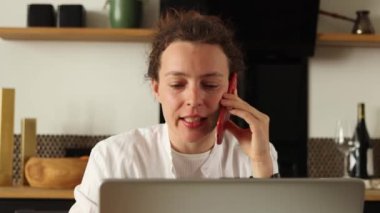 Güzel genç bayan müşteri internet sitesinden sipariş teslimatı konseptini doğrulamak için cep telefonu görüşmesi yapıyor. Telefonda dizüstü bilgisayara bakarak mutfakta otururken, beyaz elbiseler giyerken..