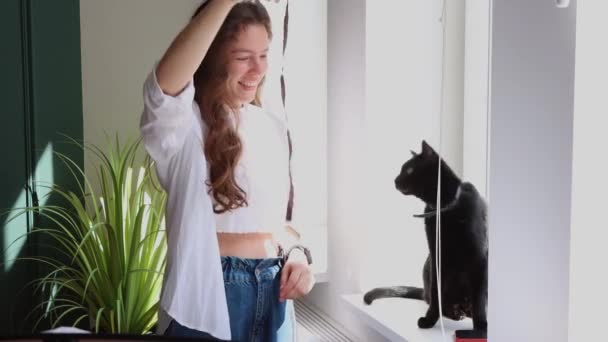 幸せな美しい女性は彼女の黒い猫と遊ぶ リボンを振って 自宅でウィンドウの近くに立って 白いシャツを着ている 素敵な子猫と一緒に晴れた週末の時間を楽しんでいるオーナー 愛らしい国産ペットのコンセプト — ストック動画