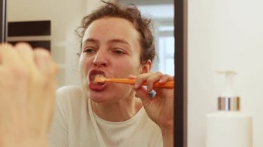 Turuncu diş fırçası tutan, dişlerini fırçalayan genç esmer kadın aynaya bakıyor. Her sabah banyoda dişçi muayenesi, yemek yedikten sonra ağız temizleme ya da kahve içme.