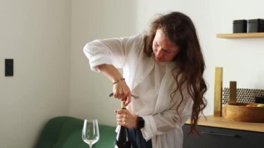 Young brunette curly woman with effort trying to open bottle of red wine using corkscrew, standing alone on kitchen at home interior, wearing white shirt. Not enough strength for uncorking alcohol. 