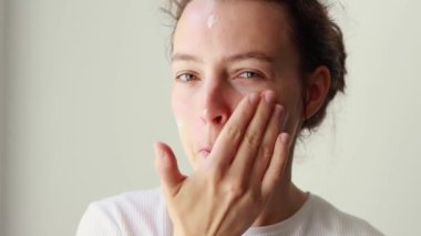 Happy attractive young caucasian woman applying skincare cream on face, looking at camera, putting soft hydrated moisturizing lifting facial creme. Beauty and healthy skin care treatment concept. 