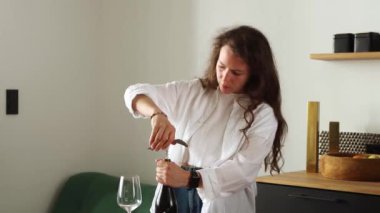 Young brunette curly woman spining corkscrew into a bottle of wine, trying to open, uncorking alcohol beverage, standing alone on kitchen at home interior, wearing white shirt. 4k footage.