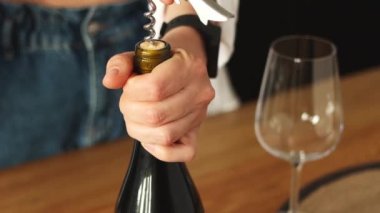 Woman hands opening wine bottle with corkscrew close up. Uncorking alcohol beverage before drinking, with wooden table and empty transparent wineglass on background indoor. 4k high quality footage.