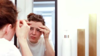 Displeased young woman looking in mirror reflection squeezing pimple on forehead, touching skin problem acne on her face in bathroom.Thinking of professional skincare products or cosmetologist meeting