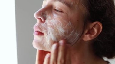 Young brunette woman washing cleaning face with organic foamy soap cleanser,back side standing in bathroom. Making massage movements. Morning beauty procedure routine, healthy skin care concept.