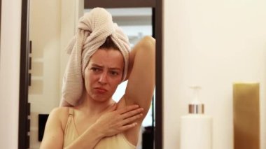 Unhappy young caucasian woman worried about black armpit with hair growing,looking in mirror reflection in bathroom at home.Feeling unsure with unwanted dark hair on the body, lacking self confidence.