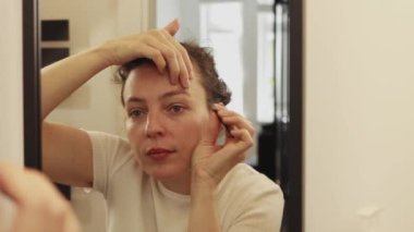 Happy attractive young caucasian woman doing her eyebrow contours sharp accurate using tweezer, looking at mirror in the bathroom.Plucking correcting thick ideal natural brows, removing hairs on face.