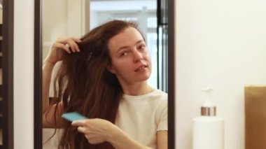 Gülümseyen genç bir kadın, esmer dalgalı saçlarını tarıyor aynaya bakıyor, sabahları banyoda saç stili yapıyor, her gün evde hazırlanıyor. Güzellik tedavi konsepti..