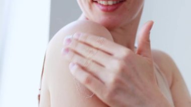 Gülümseyen genç beyaz kadın omuzlarına nemlendirici vücut losyonu sürüyor, duştan sonra günlük nemlendirici krem sürüyor. Güzellik ve sağlıklı cilt bakımı konsepti. Yavaş çekim